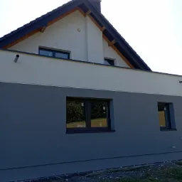 Dwukolorowa elewacja nowoczesnego domu z ciemnoszarą dolną częścią i jasnoszarą górną częścią, czarne ramy okienne, widoczny fragment dachu z czarną dachówką i drewnianymi elementami konstrukcyjnymi