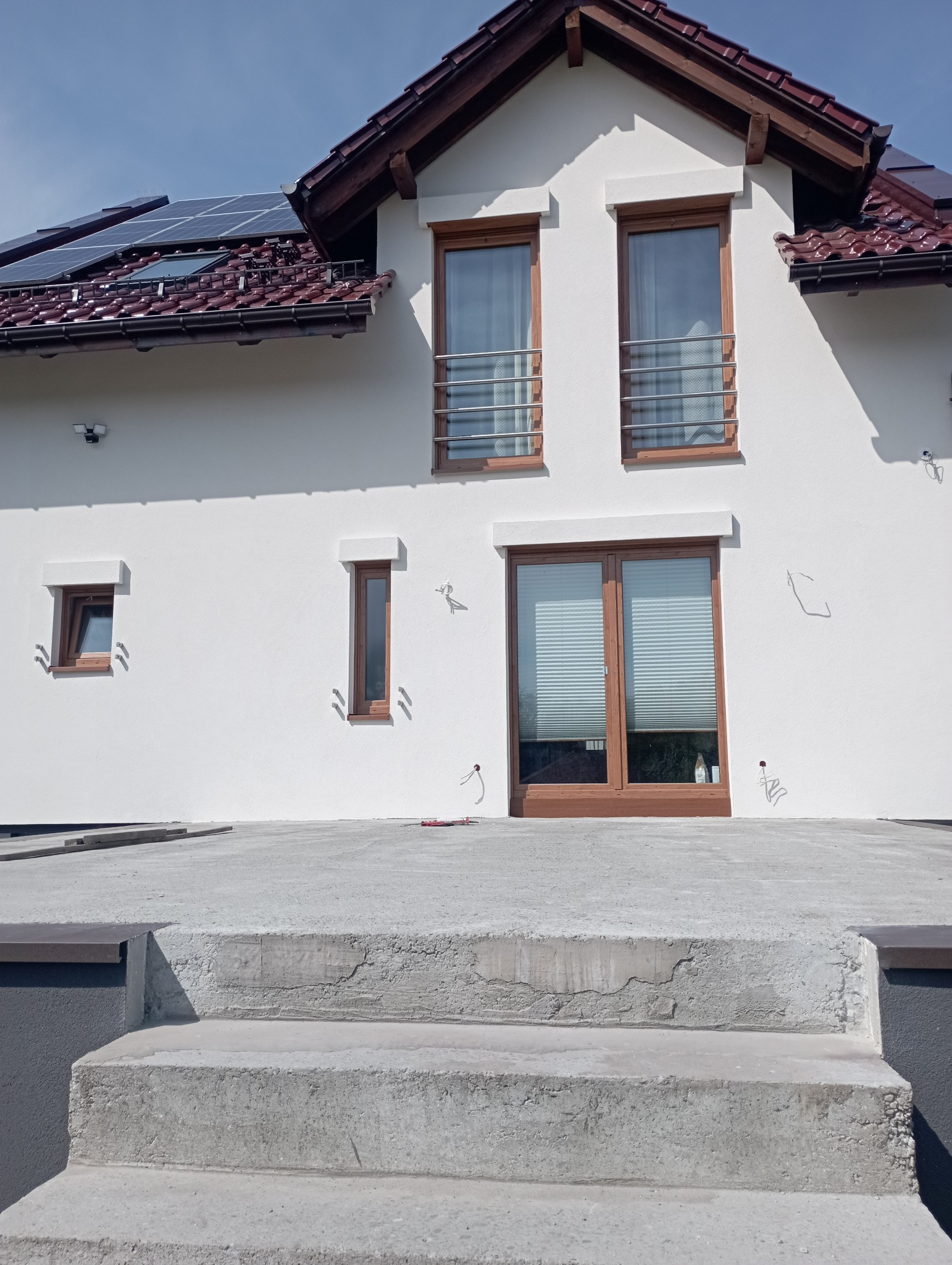 Biała elewacja domu jednorodzinnego z brązowymi oknami, widoczne panele słoneczne na dachu i betonowe schody prowadzące do wejścia.