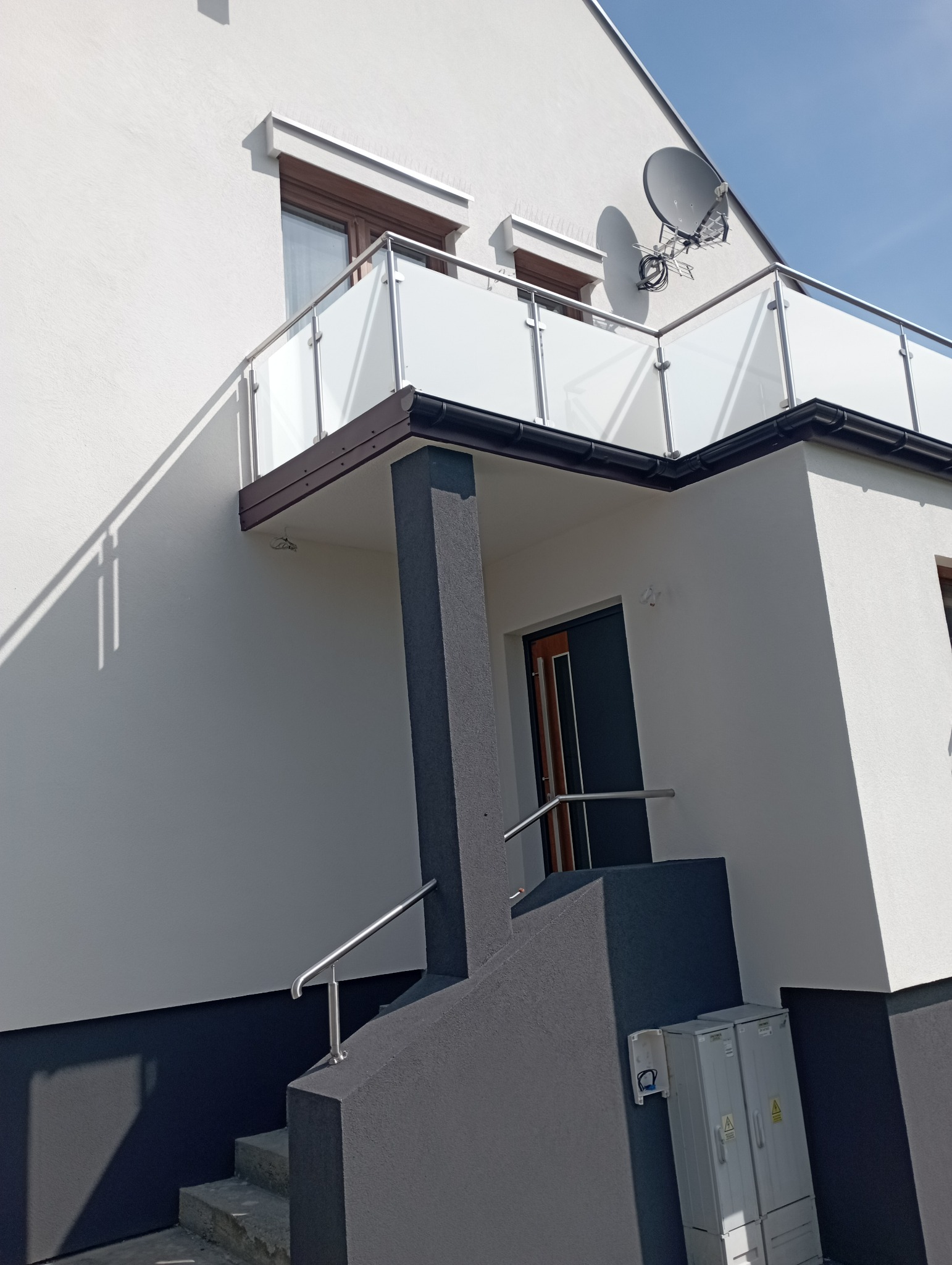 Nowoczesny dom z białą elewacją, szarymi elementami dekoracyjnymi i balkonem z szklaną balustradą, widoczna antena satelitarna na elewacji, oświetlony jasnym słońcem.