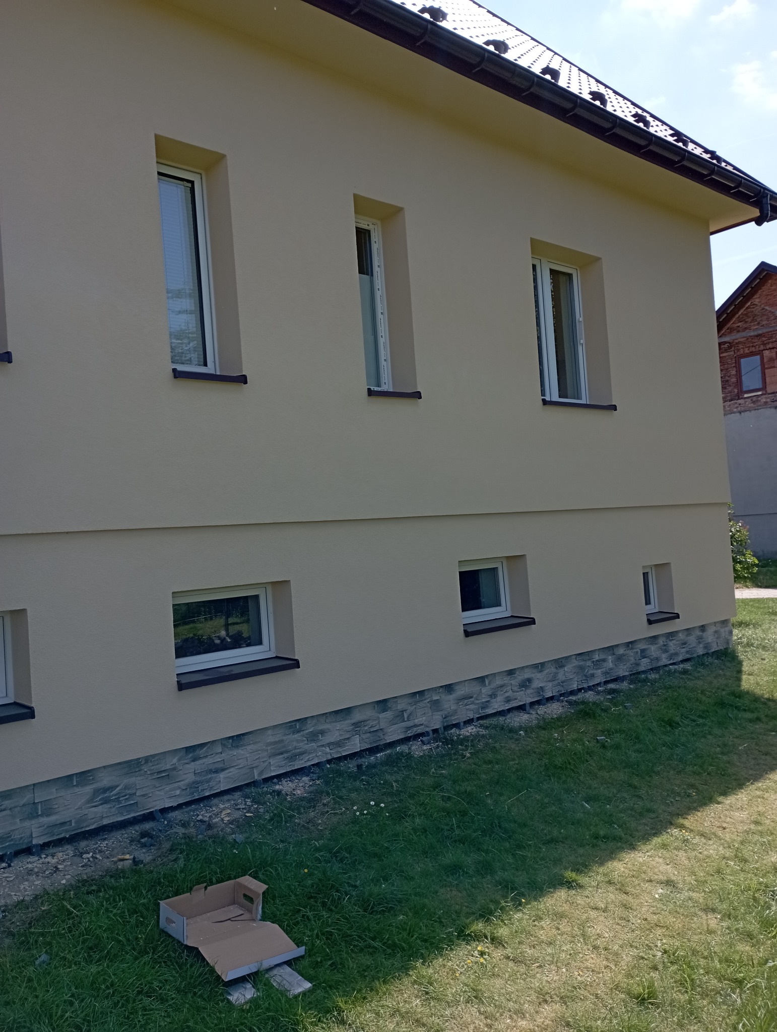 Jasnożółta elewacja domu jednorodzinnego z białymi oknami i ciemnoszarą podmurówką z imitacji kamienia, na trawniku leży rozłożony karton.