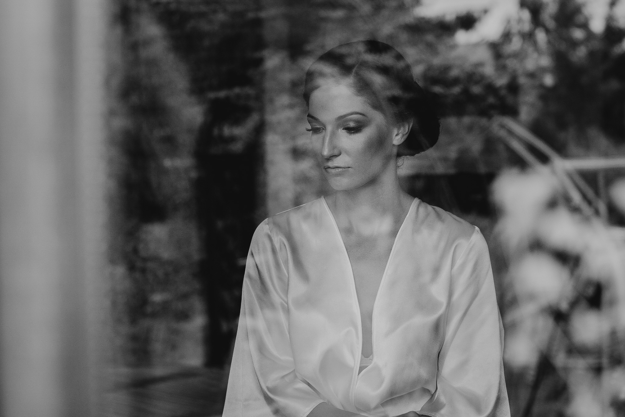 Portret kobiety w białym szlafroku, z delikatnym makijażem, spoglądającej w dół, widocznej przez rozmyte szkło okna.