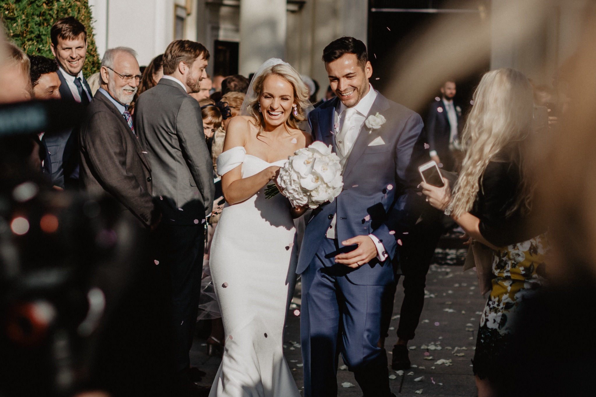 Uśmiechnięta para młoda w otoczeniu gości weselnych obsypywana płatkami kwiatów przed budynkiem.