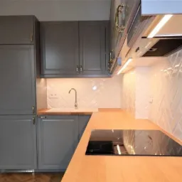 Szara zabudowa kuchenna z frezowanymi frontami, drewniany blat i białe płytki ułożone w jodełkę, oświetlenie podszafkowe.