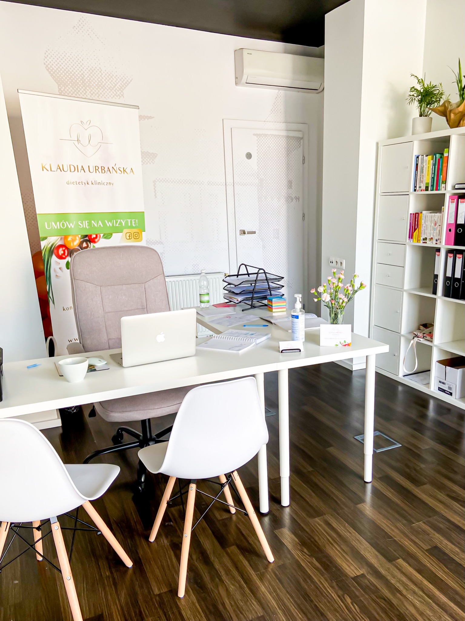 Jasne biuro z biurkiem, laptopem, krzesłami Eames, kwiatami, bannerem reklamowym i regałem z książkami, prezentujące gabinet.