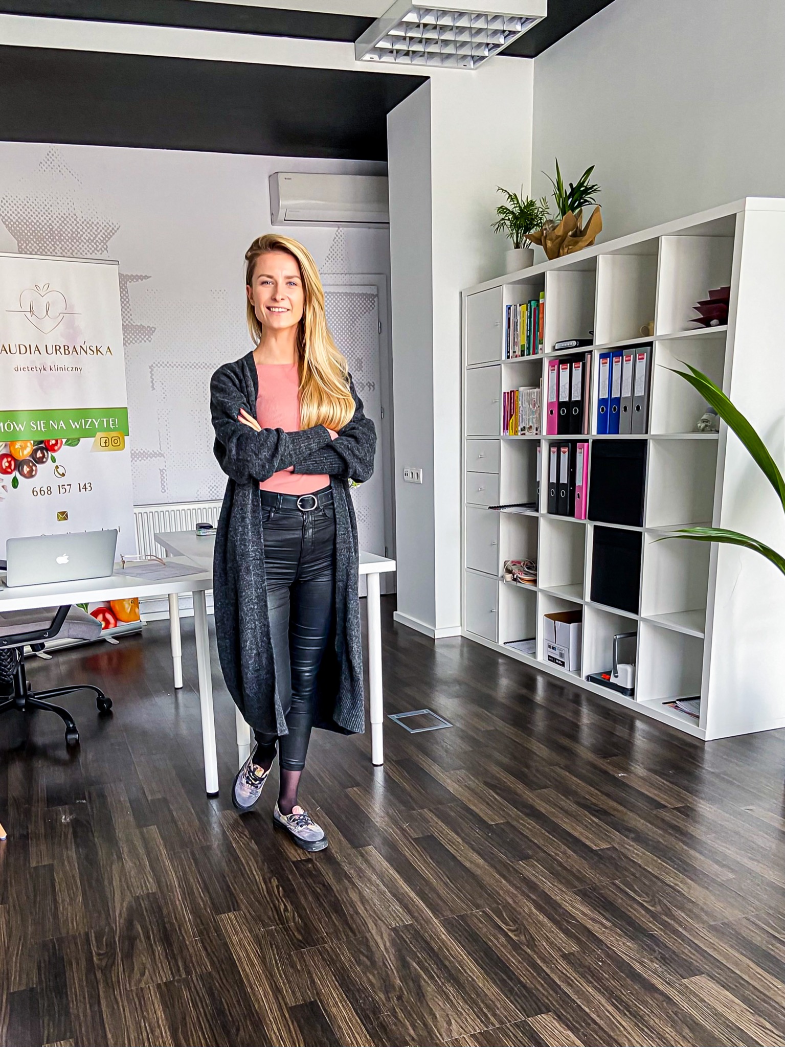 Kobieta z blond włosami, ubrana w długi kardigan i skórzane spodnie, stoi z założonymi rękami w jasnym biurze z białymi meblami i drewnianą podłogą, za nią widoczny baner reklamowy z logo...