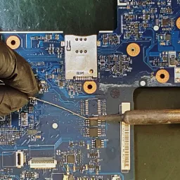 Czyszczenie płyty głównej laptopa za pomocą lutownicy i drutu przez osobę w czarnych rękawicach ochronnych.