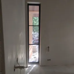 Świeżo otynkowana ściana wewnętrzna z wąskim, pionowym oknem w czarnej ramie, przez które widać drzewa i materiały budowlane na zewnątrz.