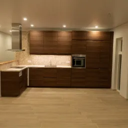 Nowoczesna, przestronna kuchnia z drewnianymi szafkami, beżową mozaiką nad blatem i minimalistycznym okapem, oświetlona punktowymi lampami sufitowymi, z płytkami podłogowymi imitującymi drewno.