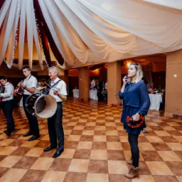Zespół muzyczny grający na weselu: akordeoniści, perkusista i wokalistka z tamburynem na parkiecie w sali z dekoracjami.