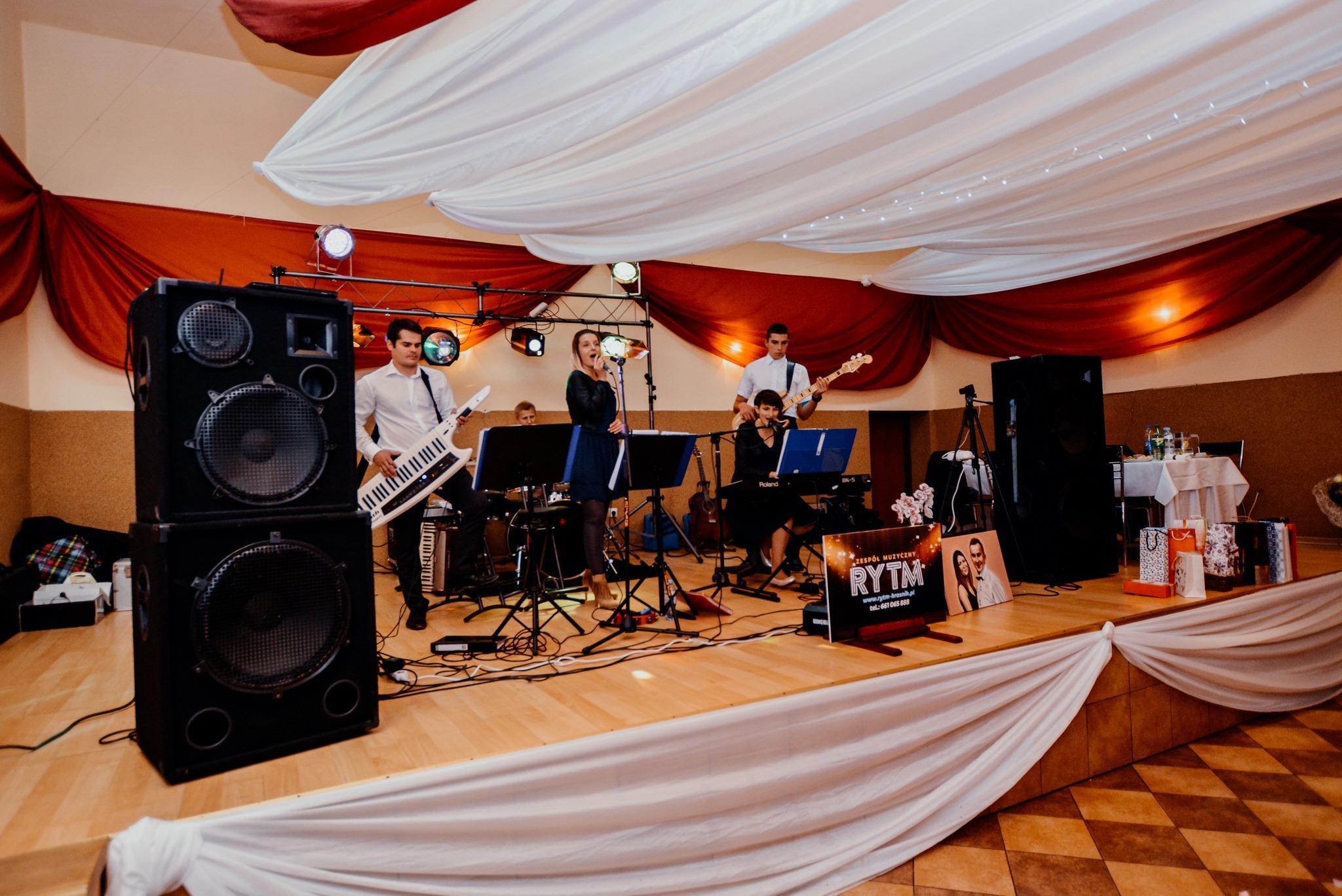 Zespół muzyczny na scenie podczas występu, wokalistka śpiewa do mikrofonu, w tle dekoracja z białych i czerwonych tkanin, widoczne instrumenty: keyboard, gitara basowa, keytar, perkusja.