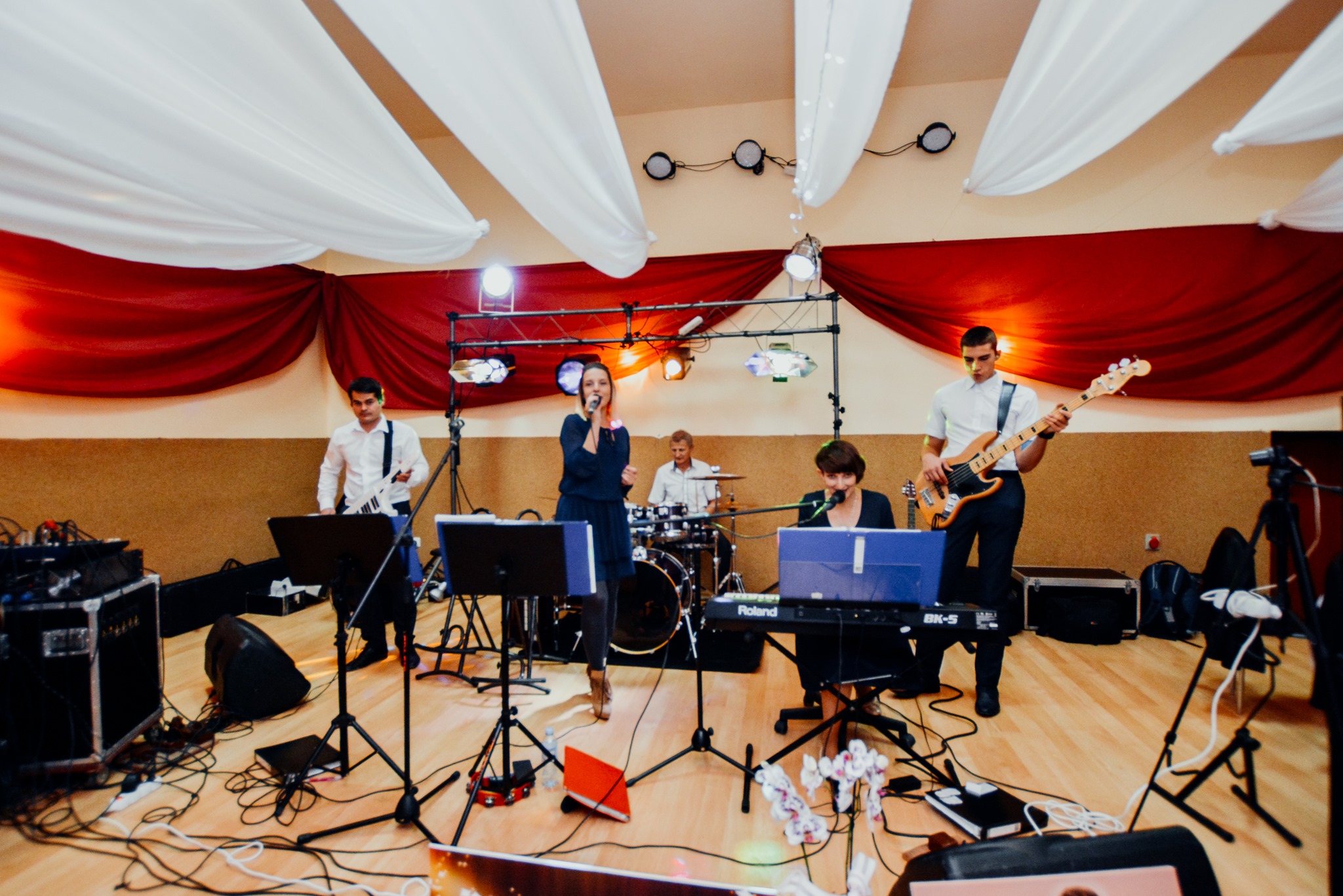 Zespół muzyczny w trakcie występu na scenie: wokalistka śpiewa do mikrofonu, klawiszowiec gra na Roland BK-5, gitarzysta basowy gra na gitarze, perkusista gra na perkusji, a saksofonista stoi...
