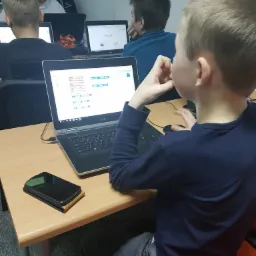 Młody chłopak siedzi przy biurku i pracuje na laptopie Dell, na ekranie widoczny interfejs programistyczny z blokami kodu, obok leży smartfon, w tle widać innych uczniów i instruktora.