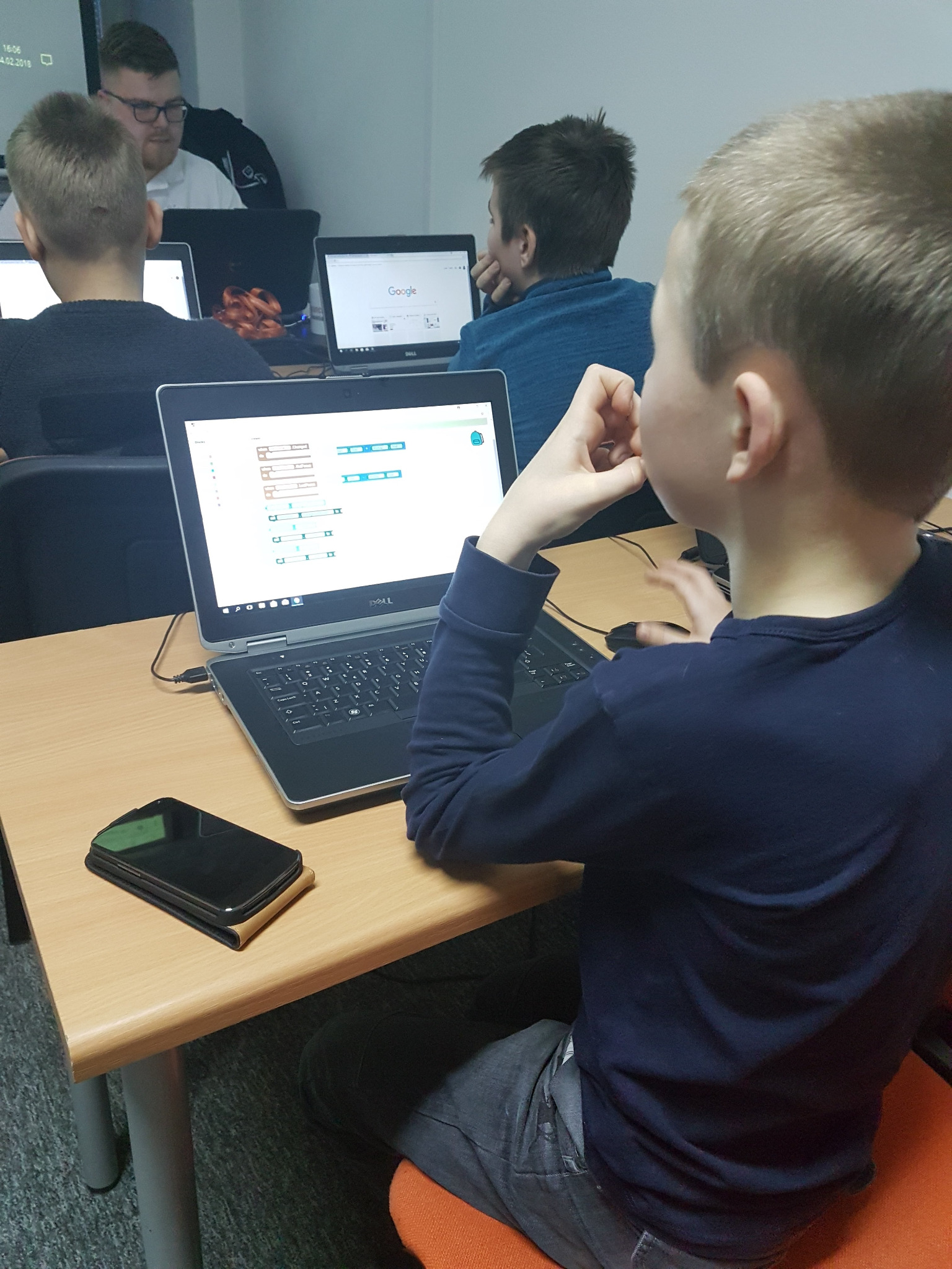 Młody chłopak siedzi przy biurku i pracuje na laptopie Dell, na ekranie widoczny interfejs programistyczny z blokami kodu, obok leży smartfon, w tle widać innych uczniów i instruktora.