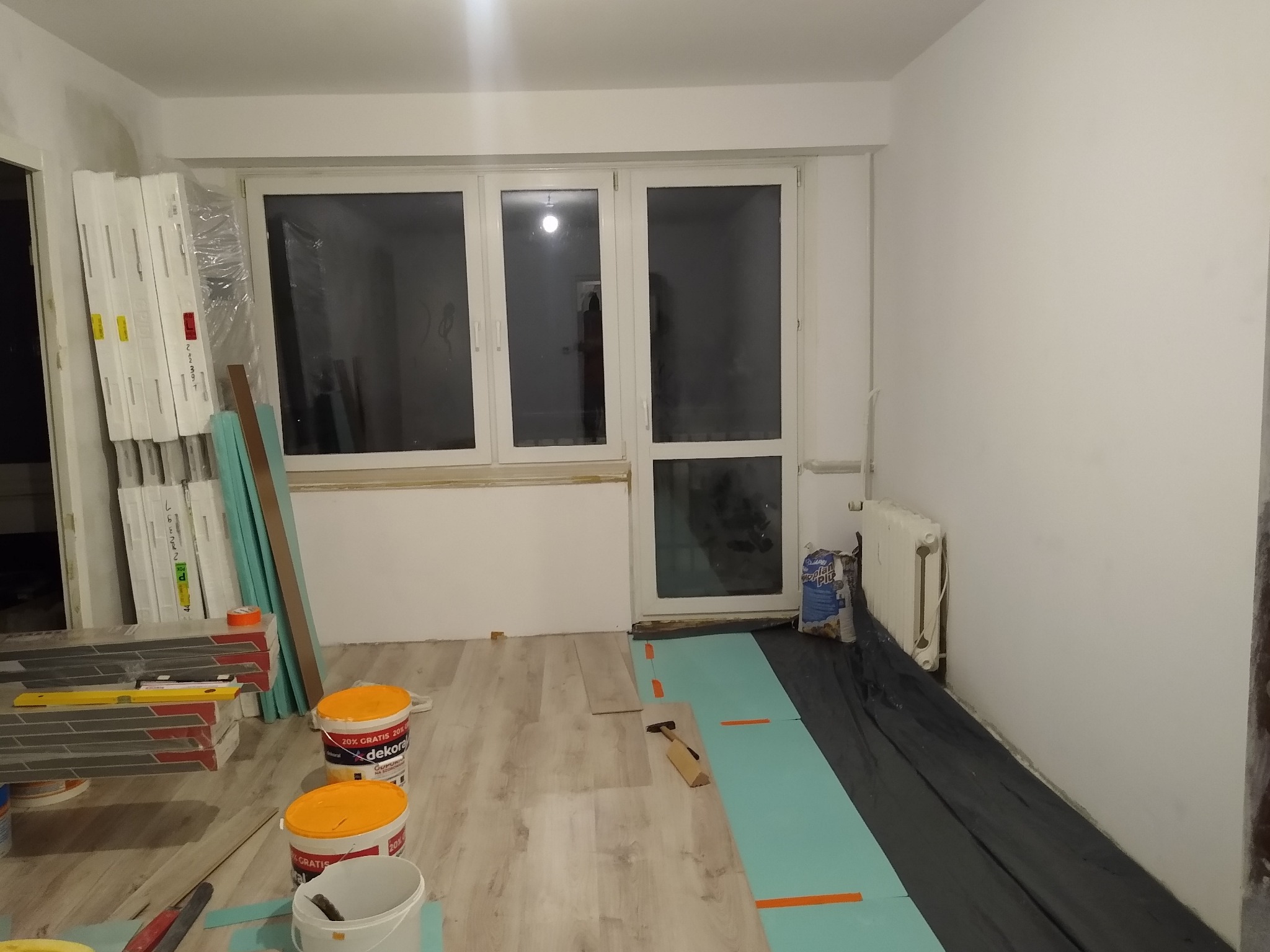 Układanie paneli podłogowych w pokoju, widoczne materiały budowlane, panele, podkład, narzędzia oraz farby w wiadrach.