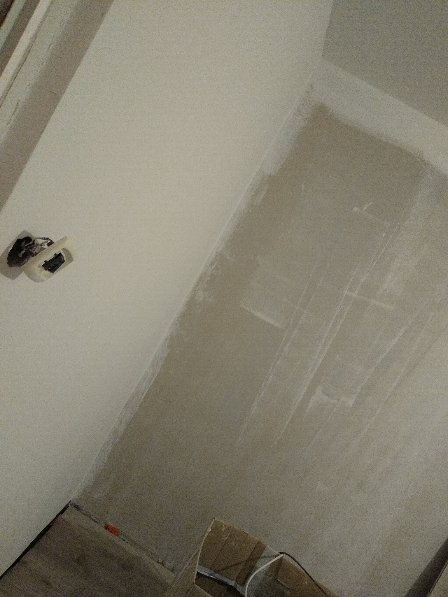 Ściana w trakcie szpachlowania z widocznym gniazdkiem elektrycznym bez obudowy i fragmentem kartonu na podłodze.