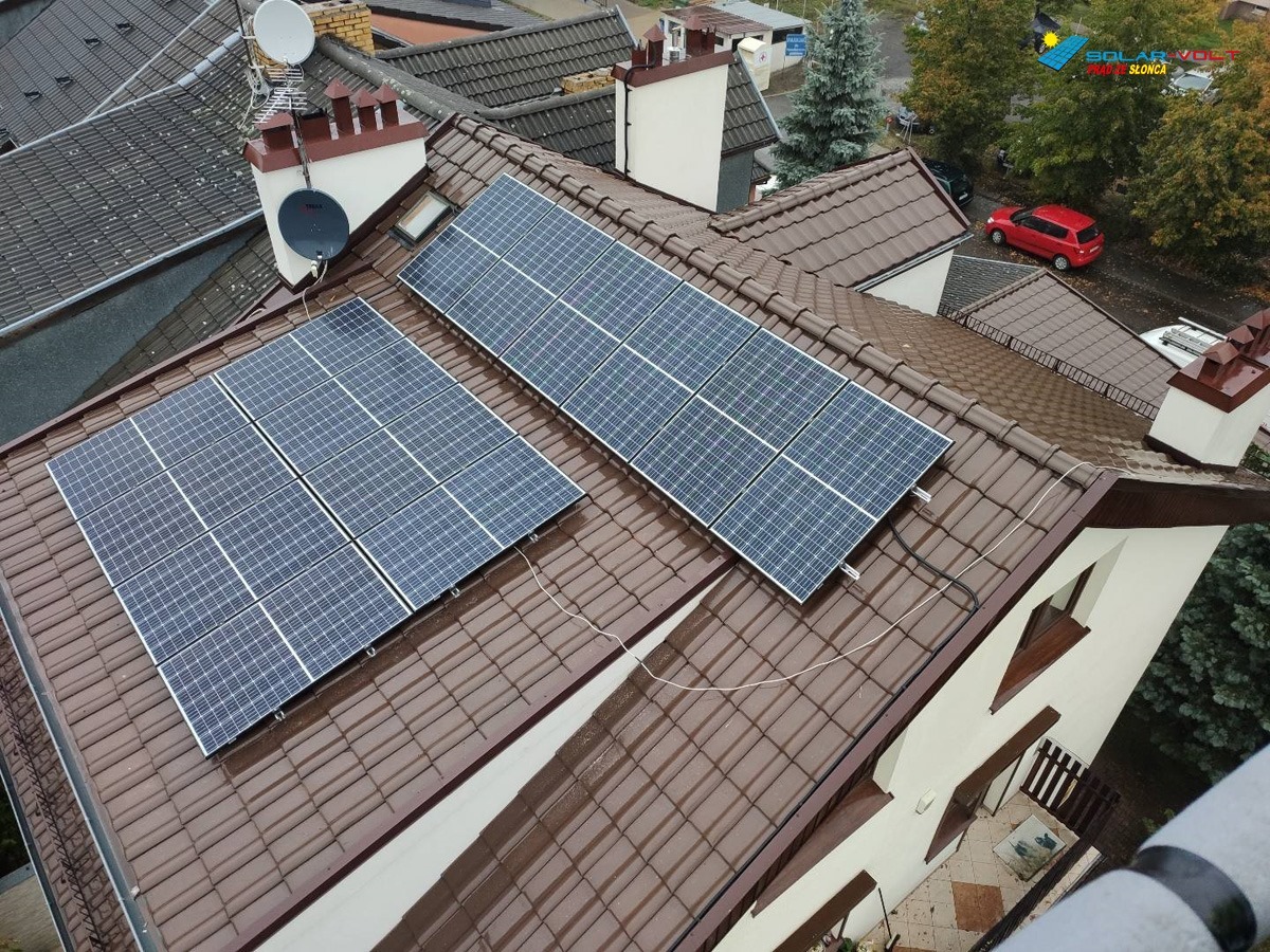 Widok z góry na brązowy dach z dwoma zestawami paneli słonecznych, anteny satelitarne na kominie i logo firmy w prawym górnym rogu.