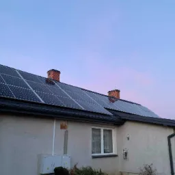 Dom jednorodzinny z panelami słonecznymi na dachu, widoczne dwa kominy z cegły i skrzynka elektryczna na elewacji budynku.
