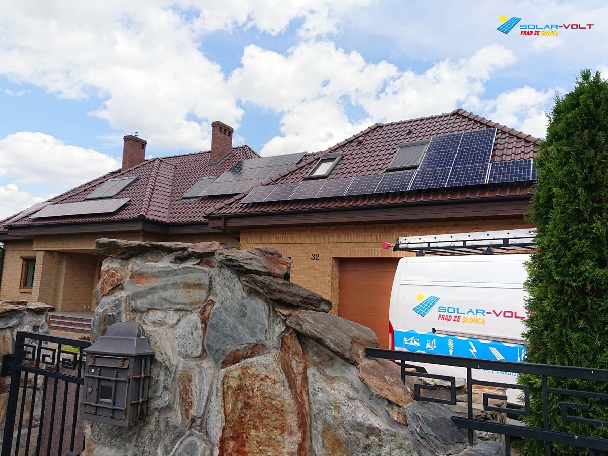 Dom jednorodzinny z panelami słonecznymi na dachu, widoczne zza kamiennego ogrodzenia i częściowo zasłonięte przez samochód serwisowy z logo firmy.