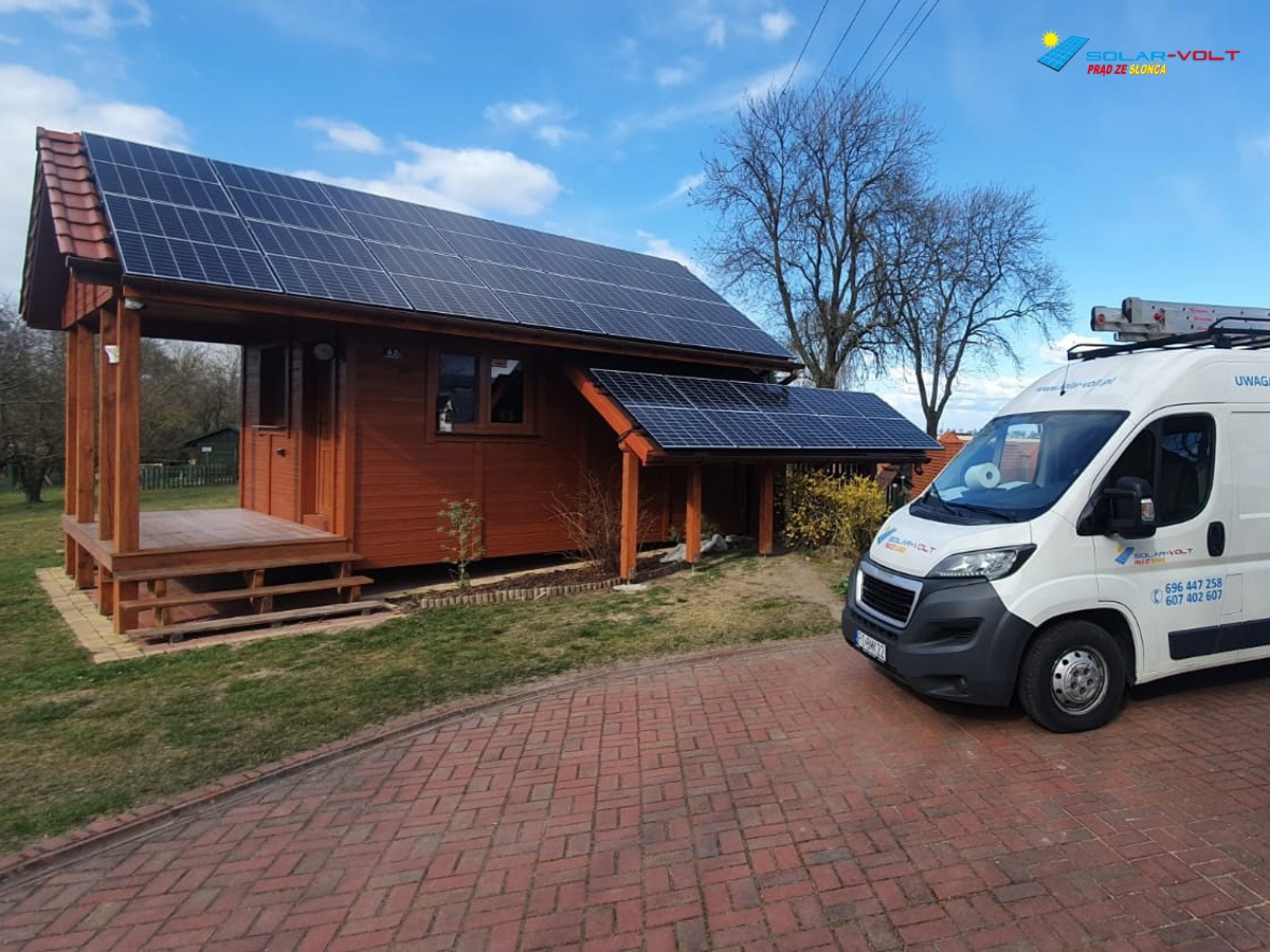 Drewniany domek z panelami słonecznymi na dachu, obok biały van z logo firmy instalującej panele. Widoczna kostka brukowa prowadząca do domku.