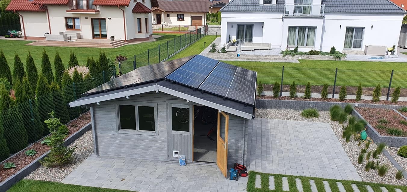 Drewniana altana ogrodowa z panelami słonecznymi na dachu, widok z góry na posesję z kostką brukową, otwartymi drzwiami, narzędziami i rowerem w środku.