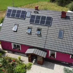 Widok z góry na dom z szarą dachówką i panelami słonecznymi, otoczony zielenią, z widocznymi oknami dachowymi i czerwonym kominem.