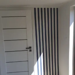 Białe drzwi wewnętrzne z poziomymi wgłębieniami i ciemną klamką na tle ściany z pionowymi, granatowo-białymi pasami.