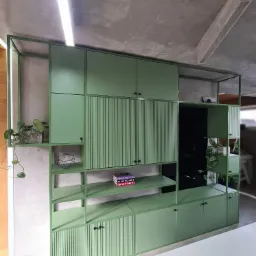 Zielony regał biurowy na wymiar z lamelami, roślinami doniczkowymi i książkami na półkach, oświetlony lampą sufitową.