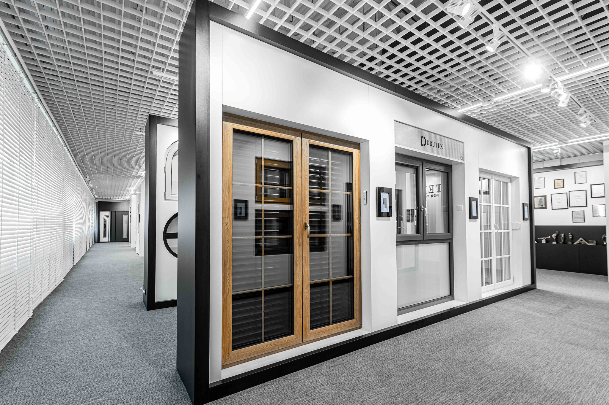 Wnętrze showroomu Drutex z ekspozycją okien i drzwi o różnych stylach i wykończeniach, w tym drewniane okno dwuskrzydłowe z wewnętrznymi żaluzjami.