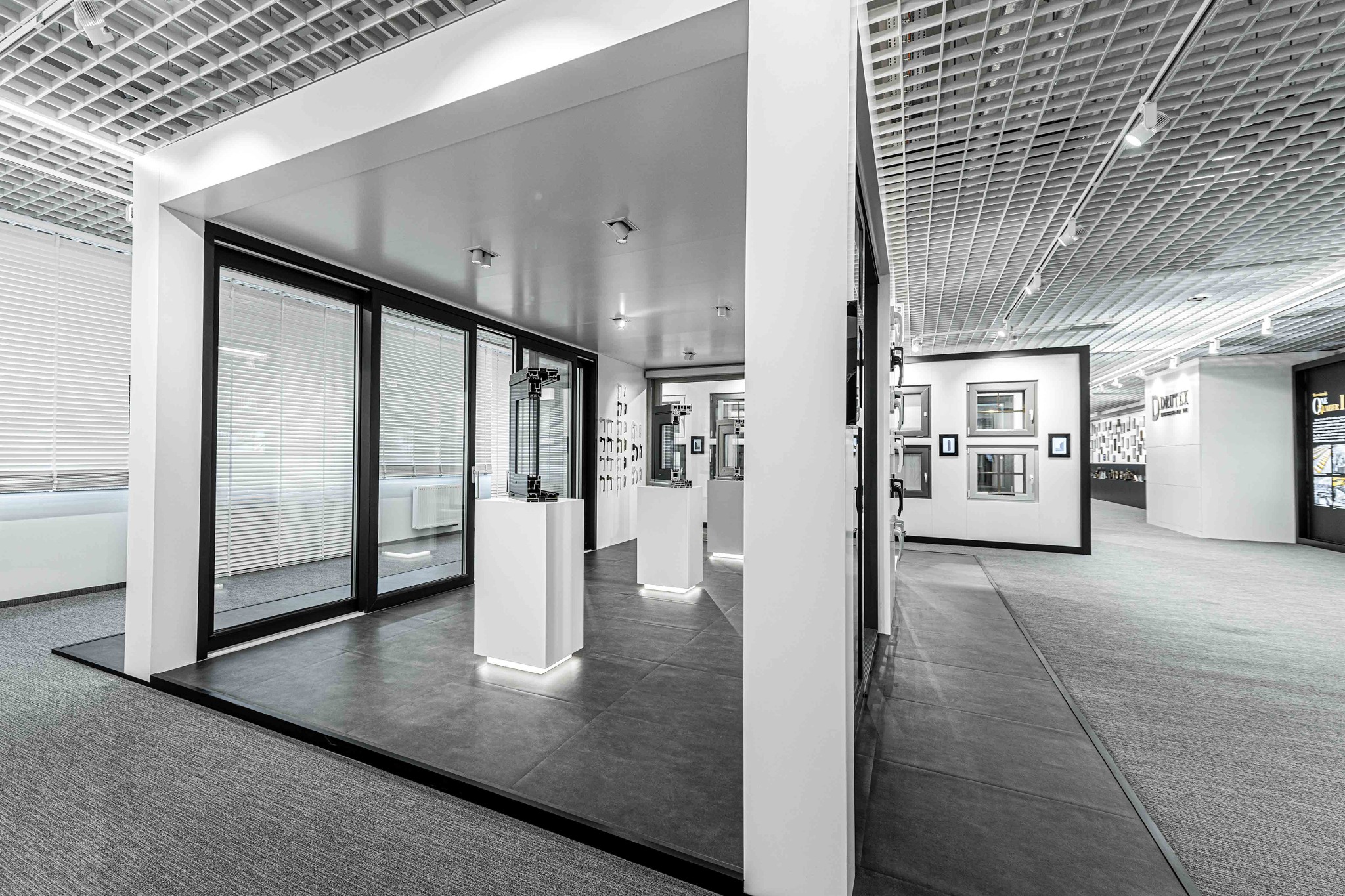 Showroom Drutex w Poznaniu prezentuje okna i drzwi balkonowe w czarnych ramach, z białymi żaluzjami, eksponowane na białych podestach z podświetleniem LED, na tle szarej podłogi i sufitu kasetonowego.