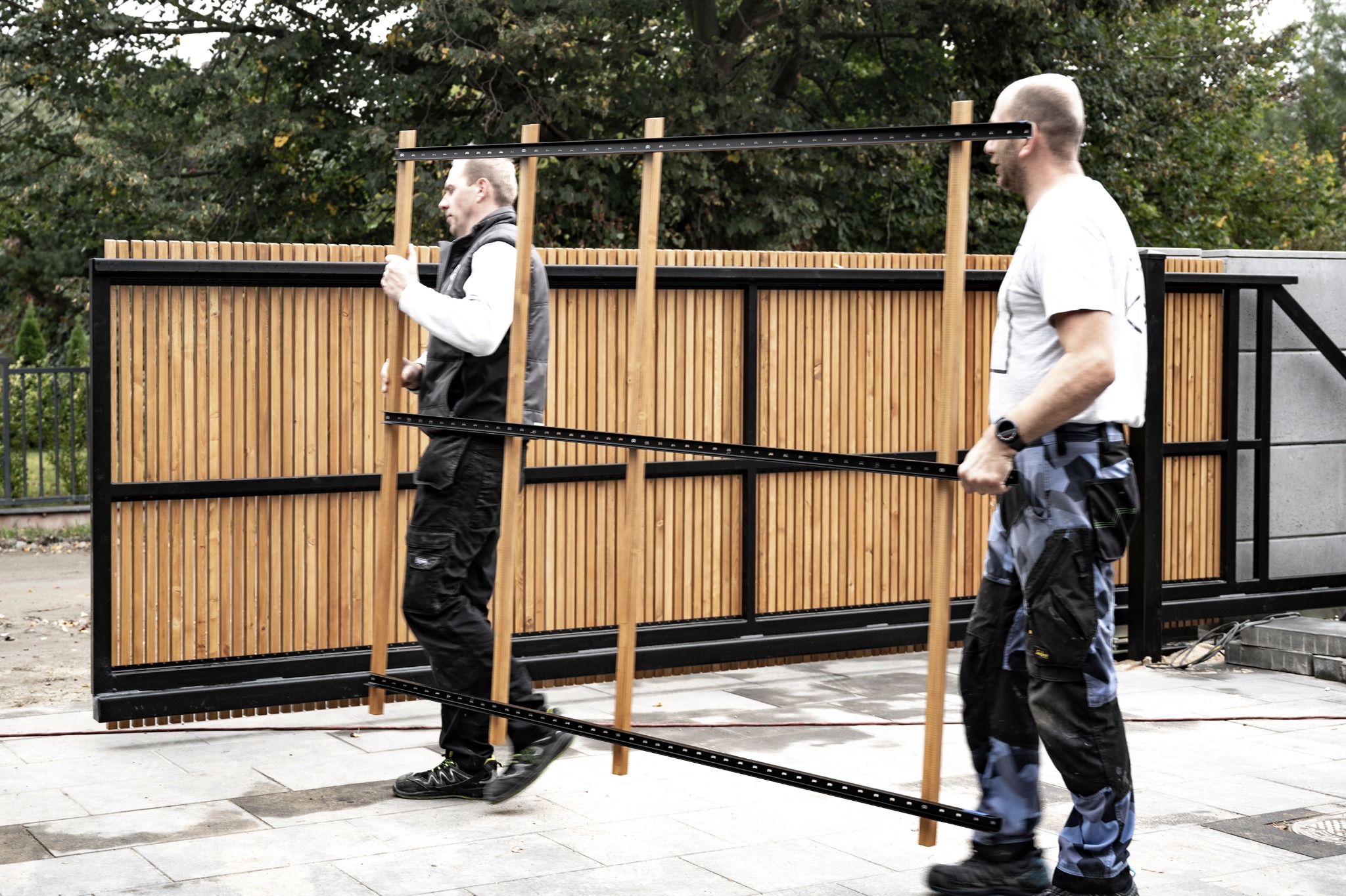 Dwóch mężczyzn w odzieży roboczej przenosi elementy drewnianego ogrodzenia na tle gotowego fragmentu płotu z pionowych desek.