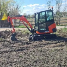 Pomarańczowa mini koparka Kubota KX027-4 na polu, zgrabione gałęzie w tle, ogrodzenie i budynki w oddali.