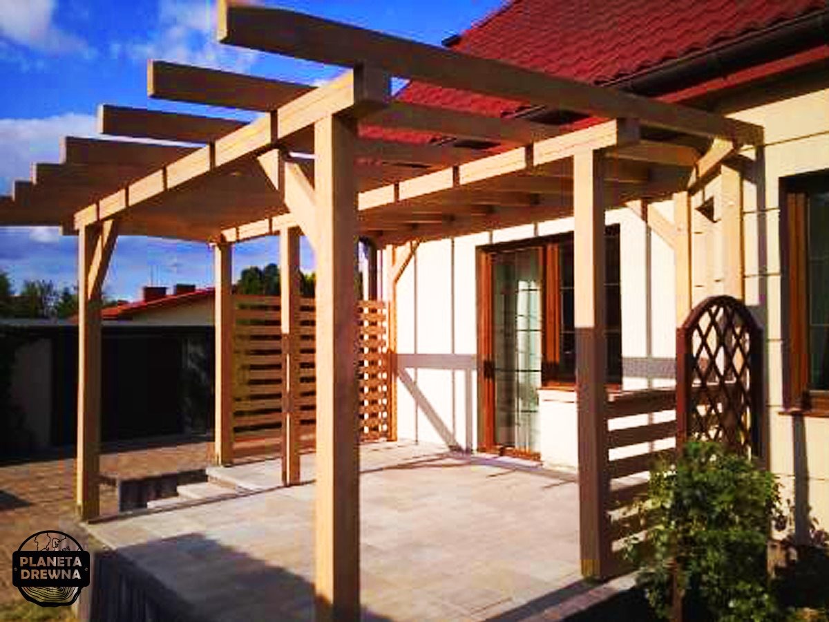 Drewniana pergola z elementami dekoracyjnymi i ażurowym panelem bocznym na tle domu z czerwoną dachówką, widoczna z poziomu tarasu wyłożonego kostką.