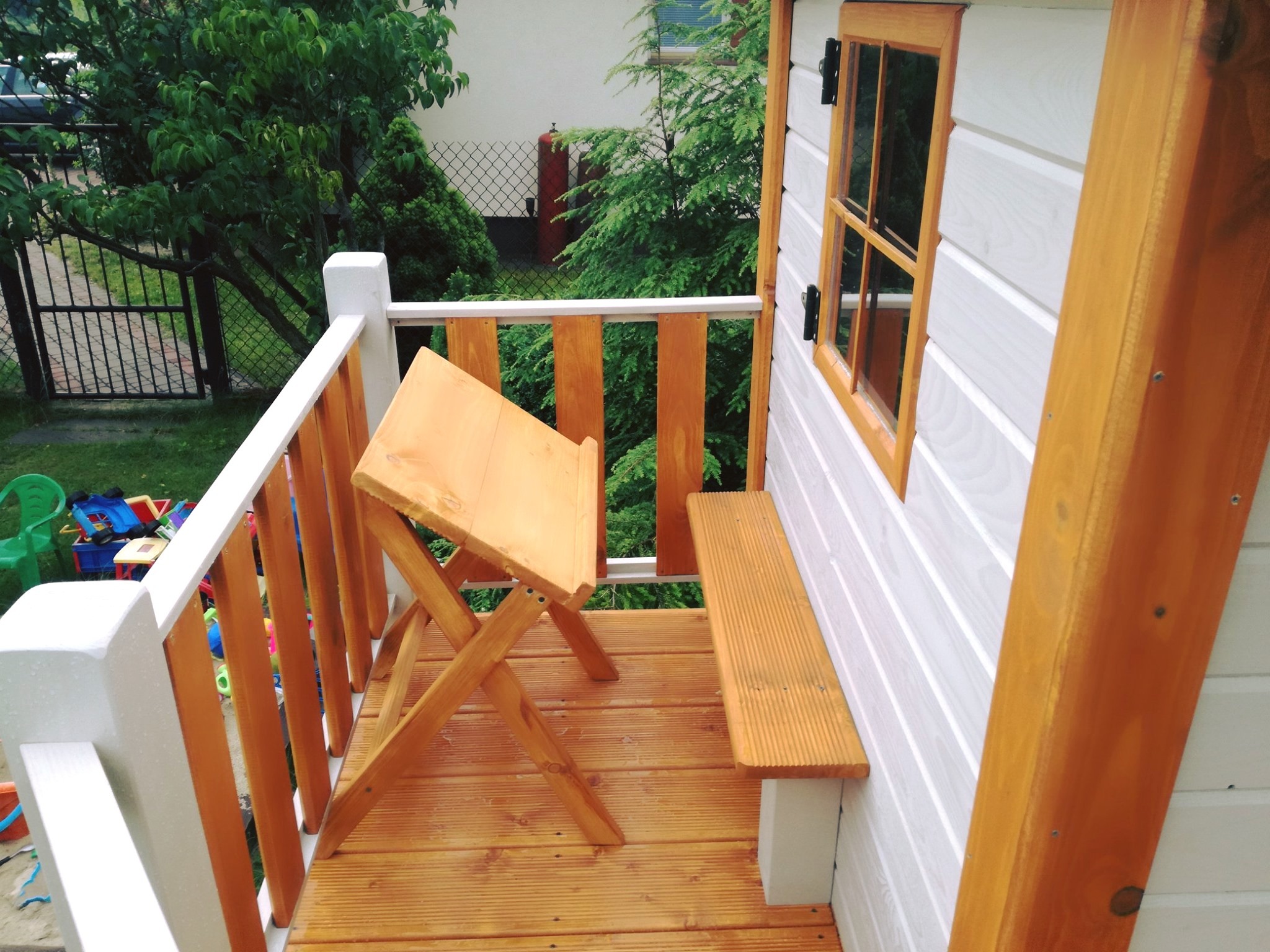 Mała drewniana altana z białymi ścianami i drewnianą podłogą, wyposażona w mały stolik składany i ławeczkę, widok z góry na zewnątrz, zabawki dziecięce w tle.