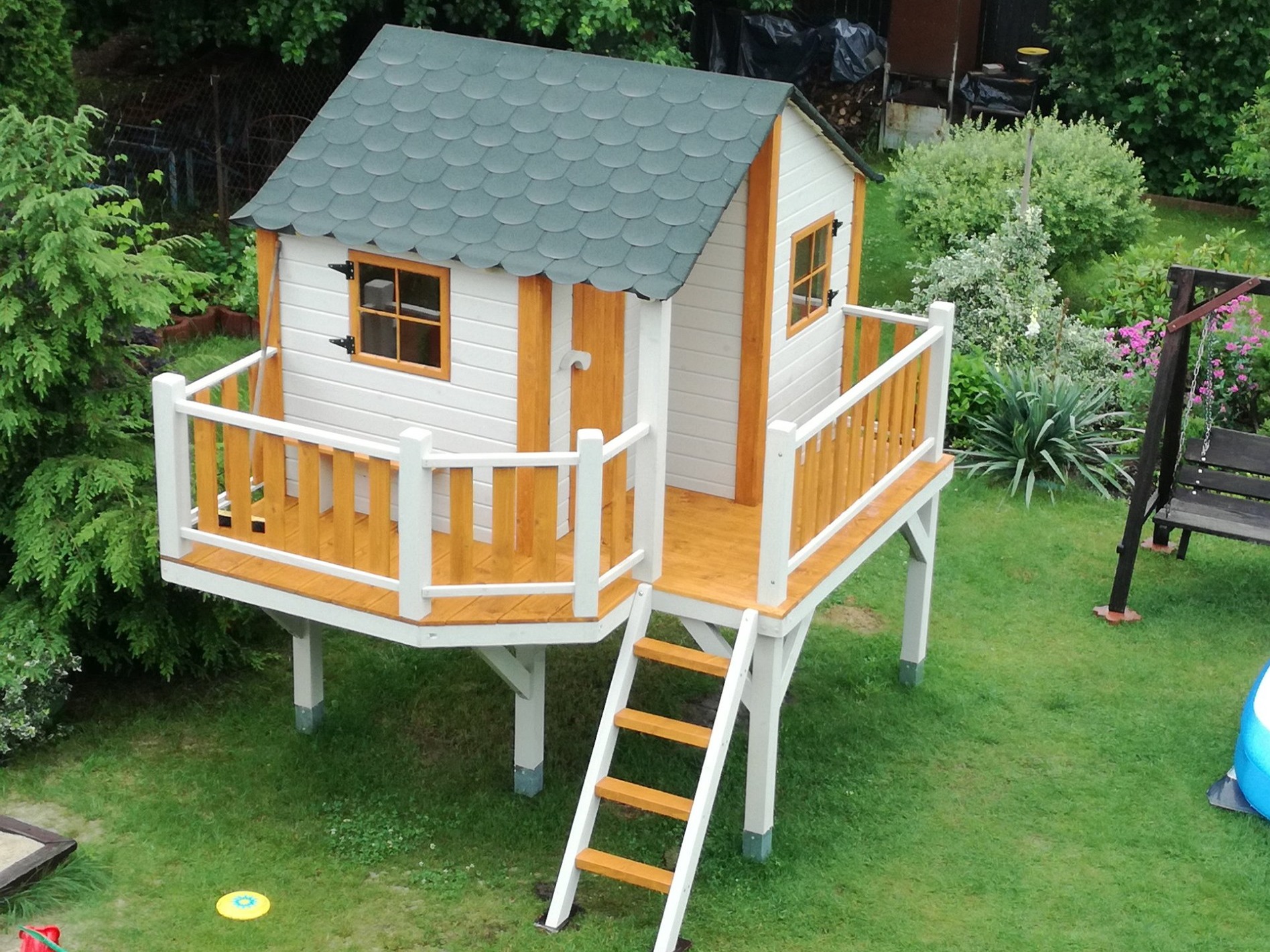 Drewniany domek dla dzieci na podwyższeniu z małym tarasem, pomalowany na biało z brązowymi akcentami, zielony dach z gontów, umieszczony w ogrodzie.