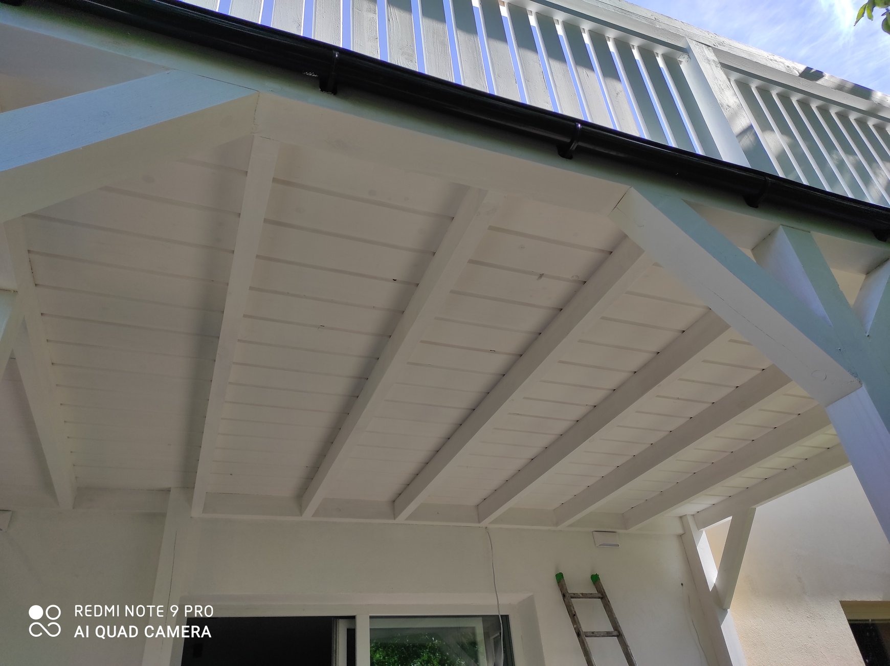 Biała drewniana konstrukcja tarasu z widocznymi belkami stropowymi i fragmentem drabiny opartą o ścianę budynku.