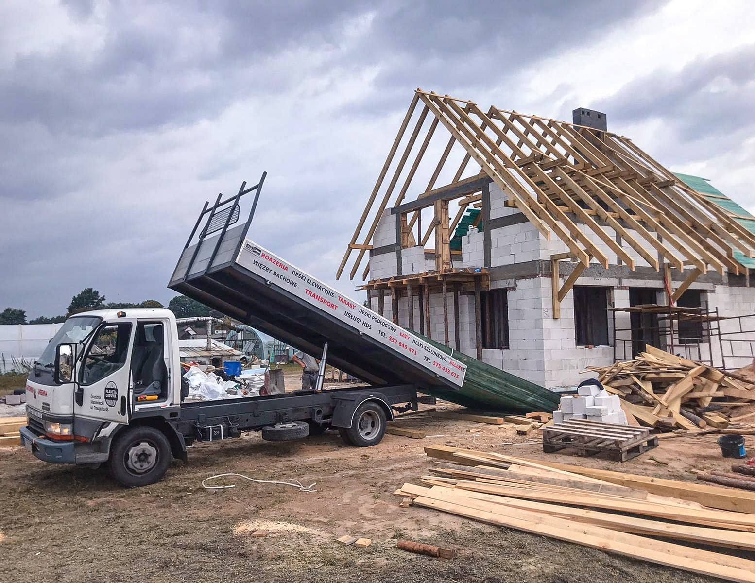 Ciężarówka z podniesioną skrzynią ładunkową, z której wysypywane są materiały budowlane obok domu w trakcie budowy, z widoczną konstrukcją dachu z drewnianych belek.