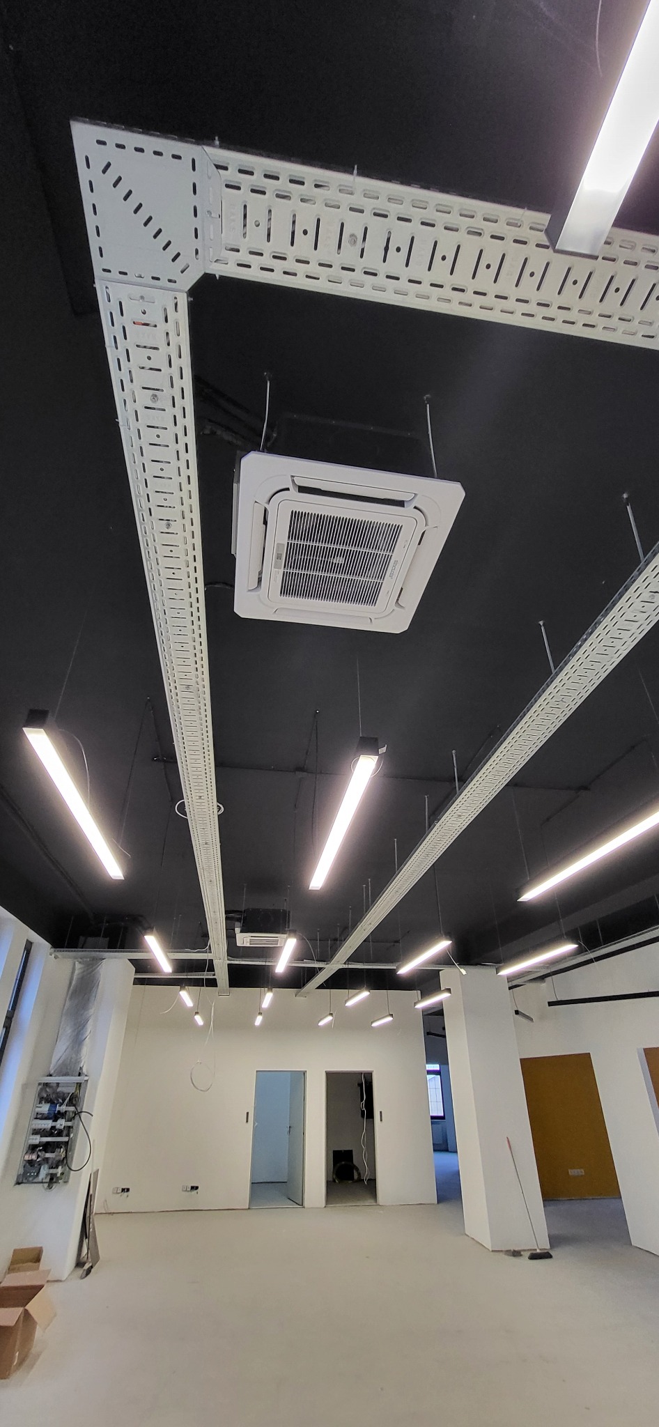 Sufit biurowy z zainstalowaną kasetonową klimatyzacją, widoczne koryta kablowe i oświetlenie liniowe. W tle surowy stan wykończenia pomieszczenia biurowego.