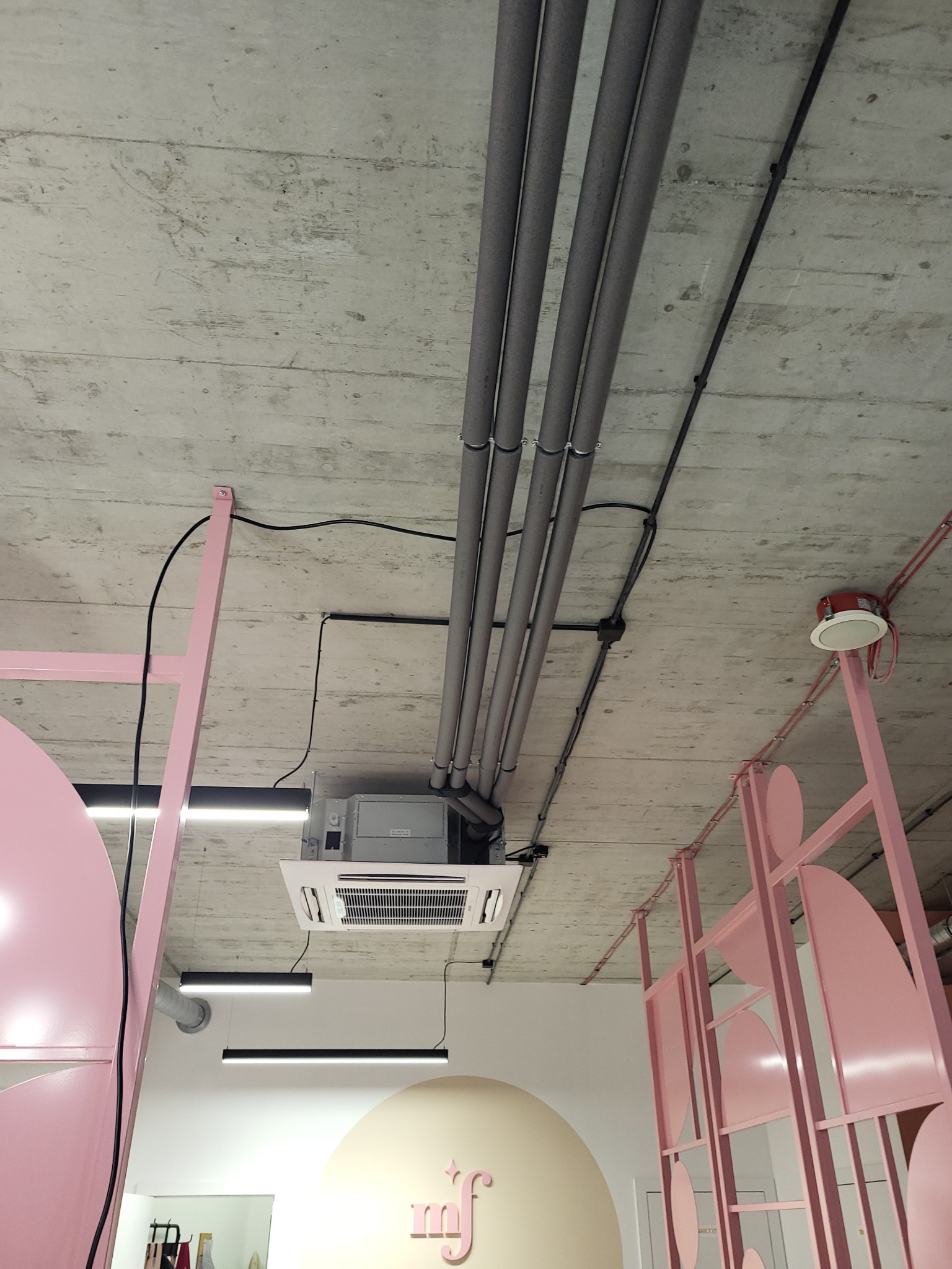 Widok na sufit w biurze z zamontowaną kasetonową klimatyzacją, widoczne rury instalacyjne, oświetlenie liniowe i dekoracyjna różowa konstrukcja.