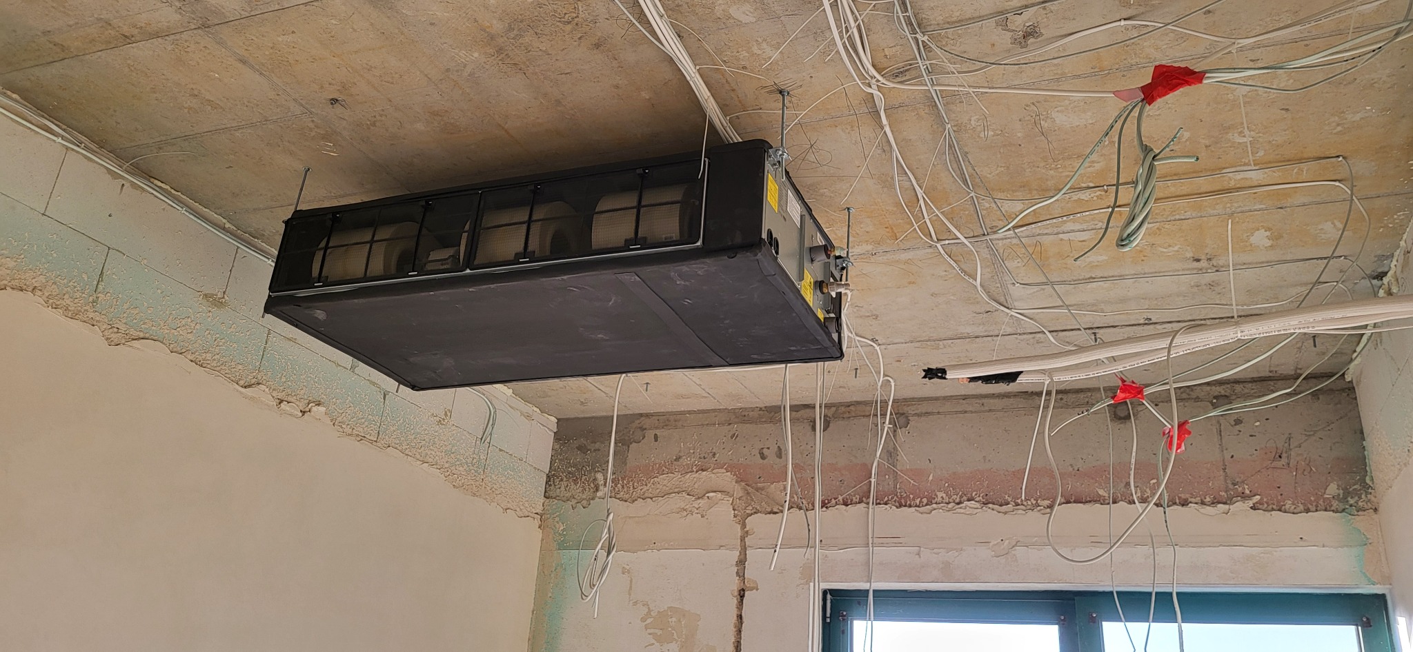 Czarna centrala klimatyzacyjna podwieszona do sufitu w surowym stanie, widoczne liczne kable i instalacje elektryczne, ściany w trakcie wykończenia.