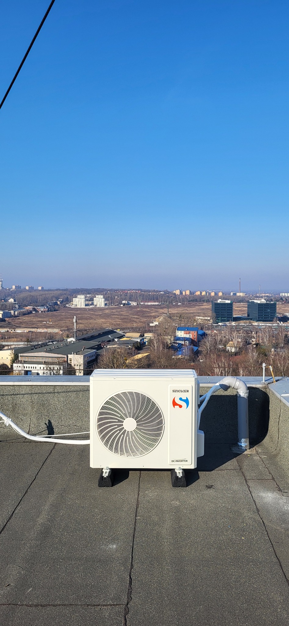 Zewnętrzna jednostka klimatyzacji marki Sinclair zamontowana na płaskim dachu z widokiem na panoramę miasta w oddali.