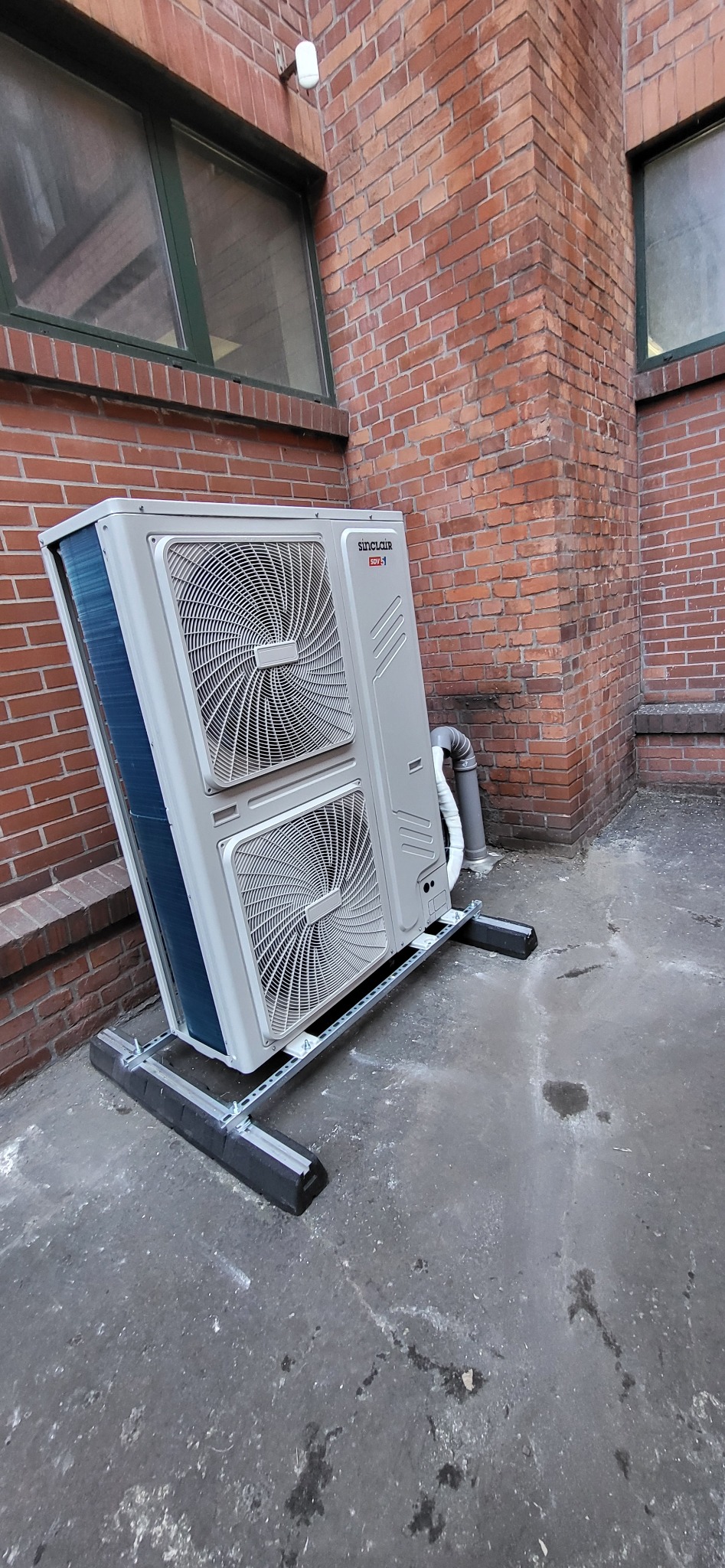 Zewnętrzna jednostka klimatyzacji Sinclair SDV z dwoma wentylatorami, zamontowana na metalowej konstrukcji na tle ceglanego budynku z oknami.