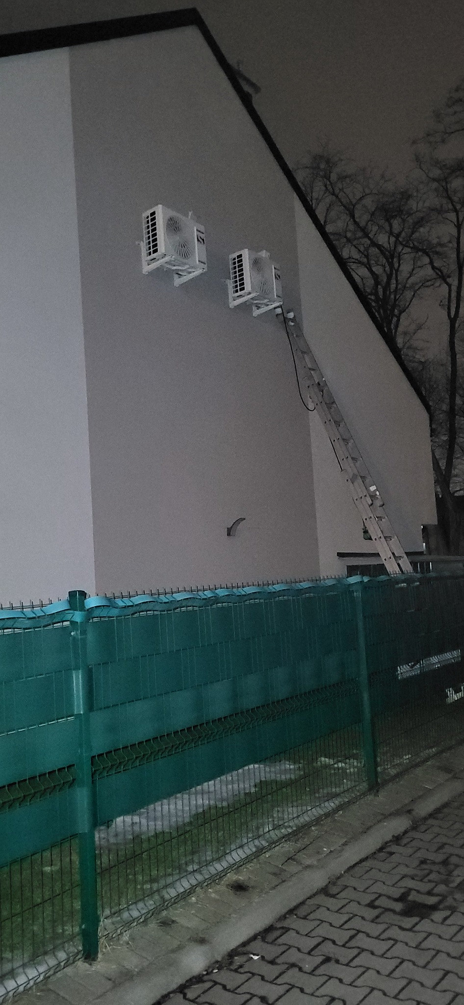Dwa klimatyzatory zamontowane na ścianie budynku, z drabiny oparta o ścianę. Widok zza zielonego ogrodzenia.