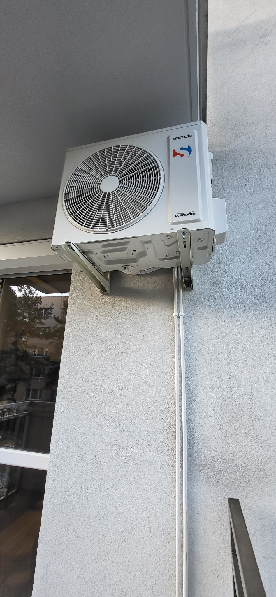 Biała jednostka zewnętrzna klimatyzacji Sinclair zamontowana na balkonie, widoczne metalowe wsporniki mocujące i białe rurki instalacyjne.