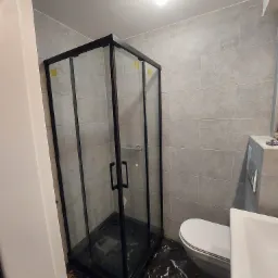 Nowoczesna kabina prysznicowa z czarną ramą i szklanymi ściankami, marmurowa podłoga, widoczna toaleta i umywalka, szare płytki na ścianach, surowa żarówka na suficie.