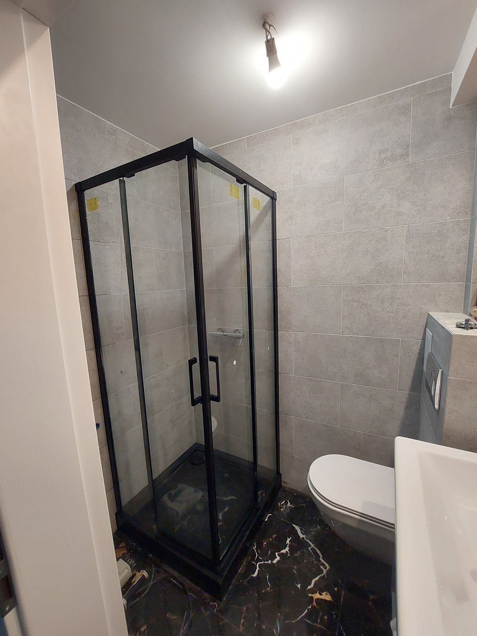 Nowoczesna kabina prysznicowa z czarną ramą i szklanymi ściankami, marmurowa podłoga, widoczna toaleta i umywalka, szare płytki na ścianach, surowa żarówka na suficie.