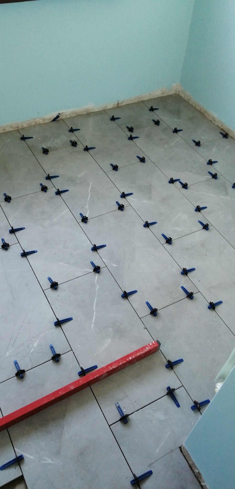 Szare płytki podłogowe w trakcie układania, z niebieskimi klipsami poziomującymi i czerwoną poziomicą, widok z góry, jasne pomieszczenie.