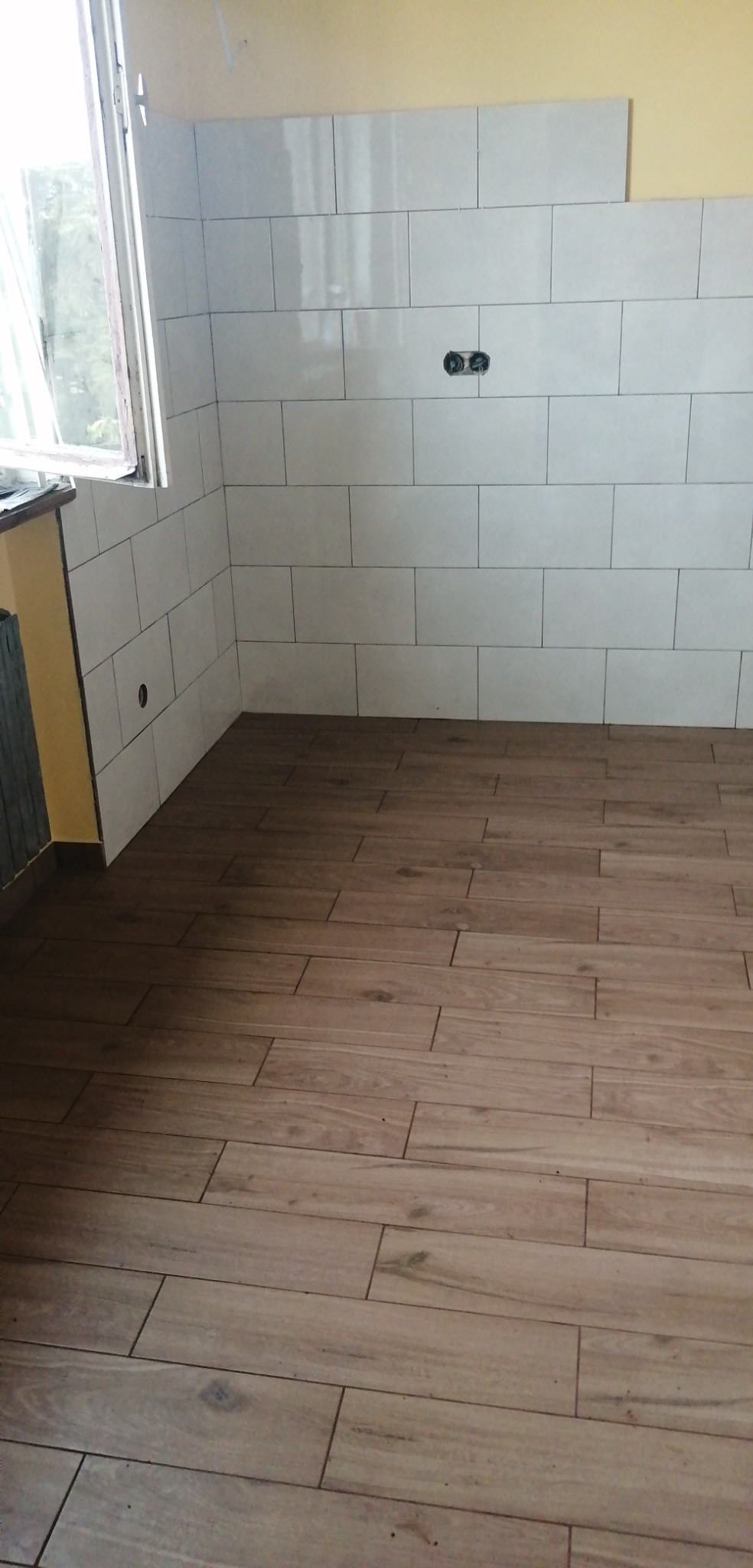 Świeżo ułożone, podłużne płytki imitujące drewno na podłodze i białe, prostokątne płytki na ścianie z widocznym gniazdkiem elektrycznym, w rogu pomieszczenia z oknem.