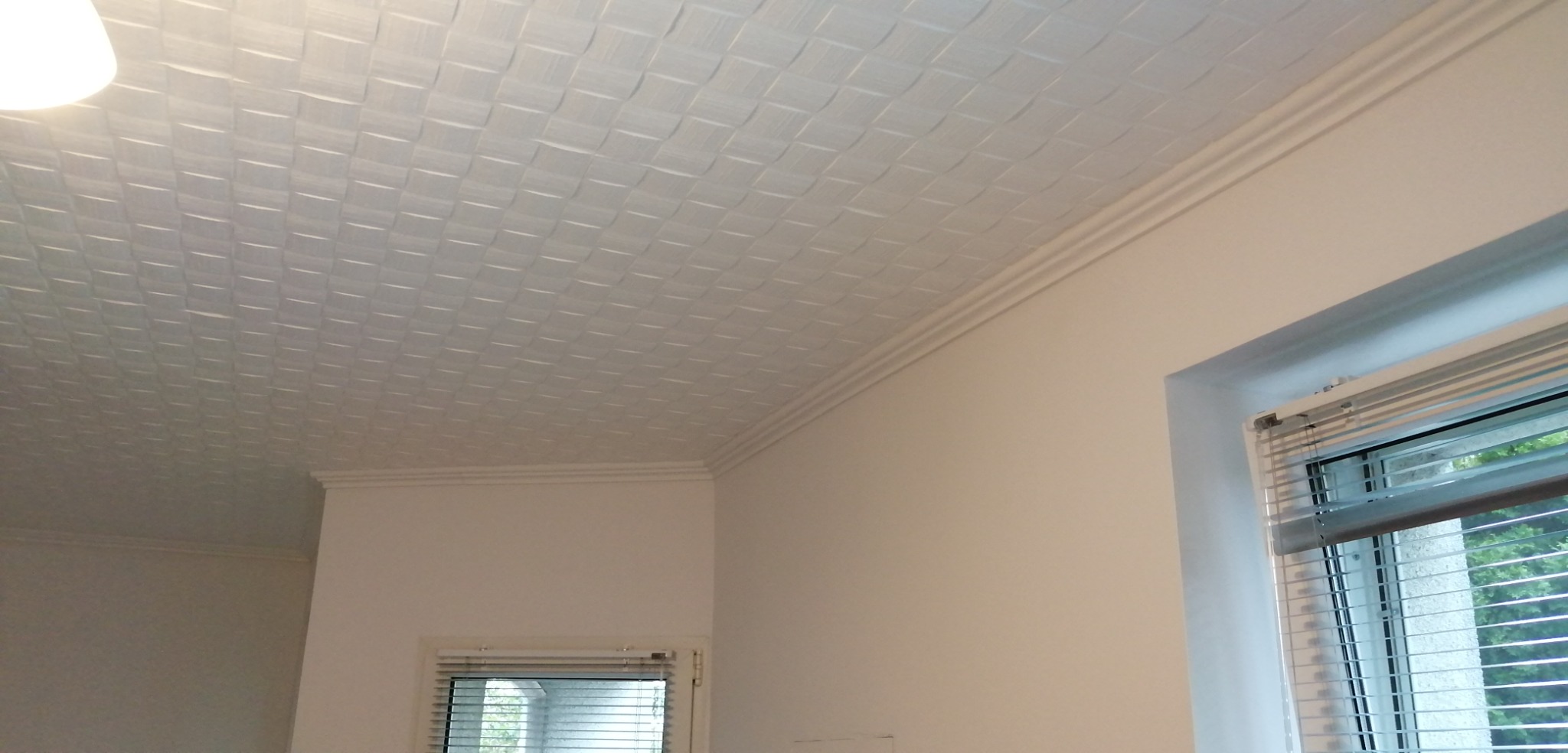 Biały sufit z dekoracyjną, kasetonową strukturą i listwami przypodłogowymi, widok z dołu, fragment okna z żaluzjami w tle.