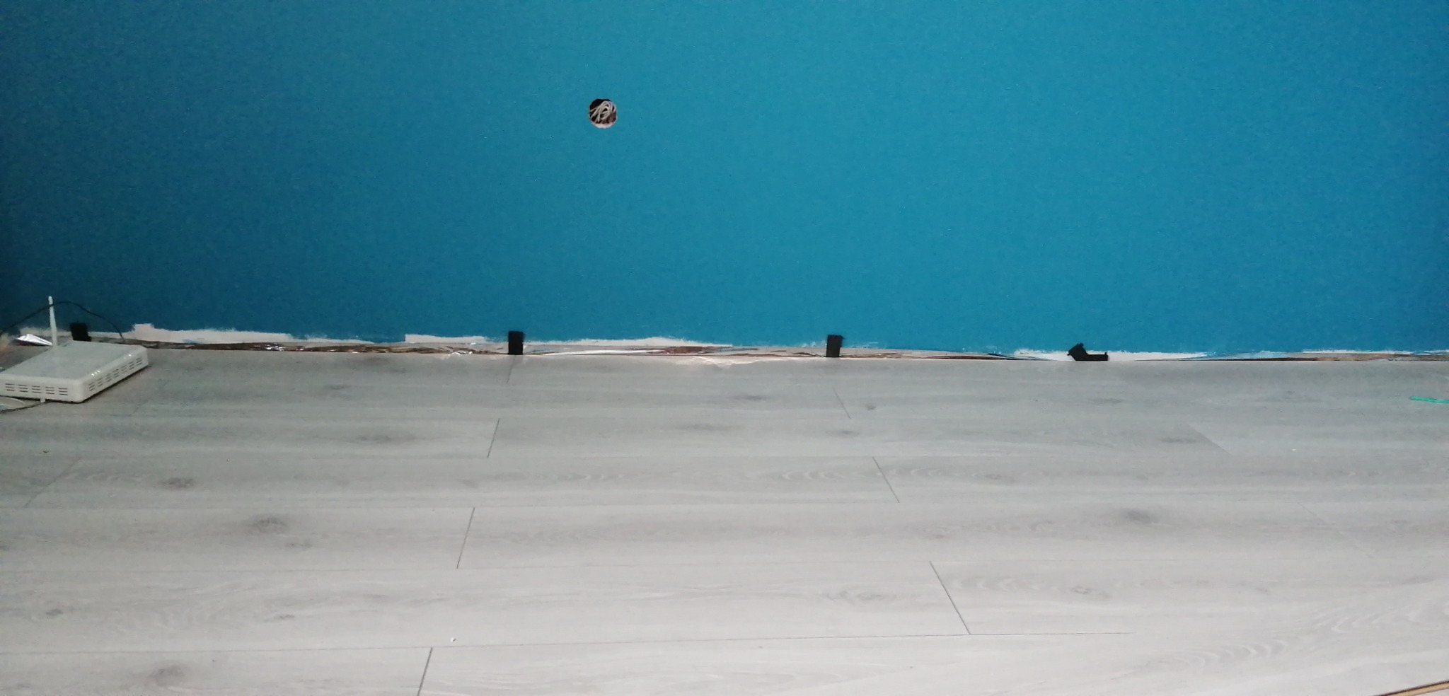 Niedokończone malowanie turkusowej ściany nad nową, jasną podłogą laminowaną, widoczny kabel w ścianie i router przy ścianie.