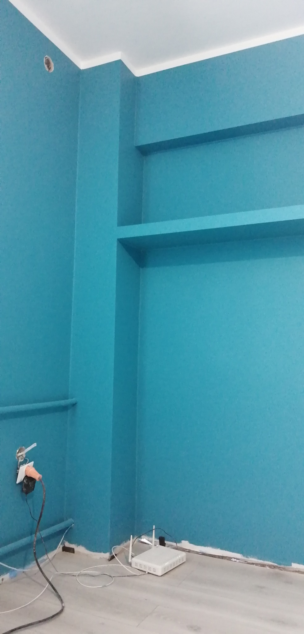 Turkusowe ściany z wnękami i wystającymi rurami, widoczny biały router i gniazdko elektryczne z podłączonym przewodem.
